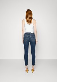 Mujer con cabello rojo vistiendo una camiseta sin mangas blanca, jeans ajustados de un azul oscuro y sandalias de tacón amarillo, de pie de espaldas frente a una pared blanca lisa.