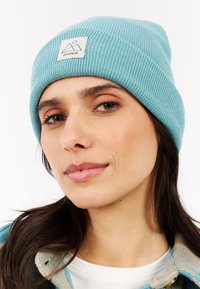 Vrouw met een lichtblauwe gebreide beanie met een "protest" logo patch, donker haar en een geruite jas over een witte top, tegen een witte achtergrond.