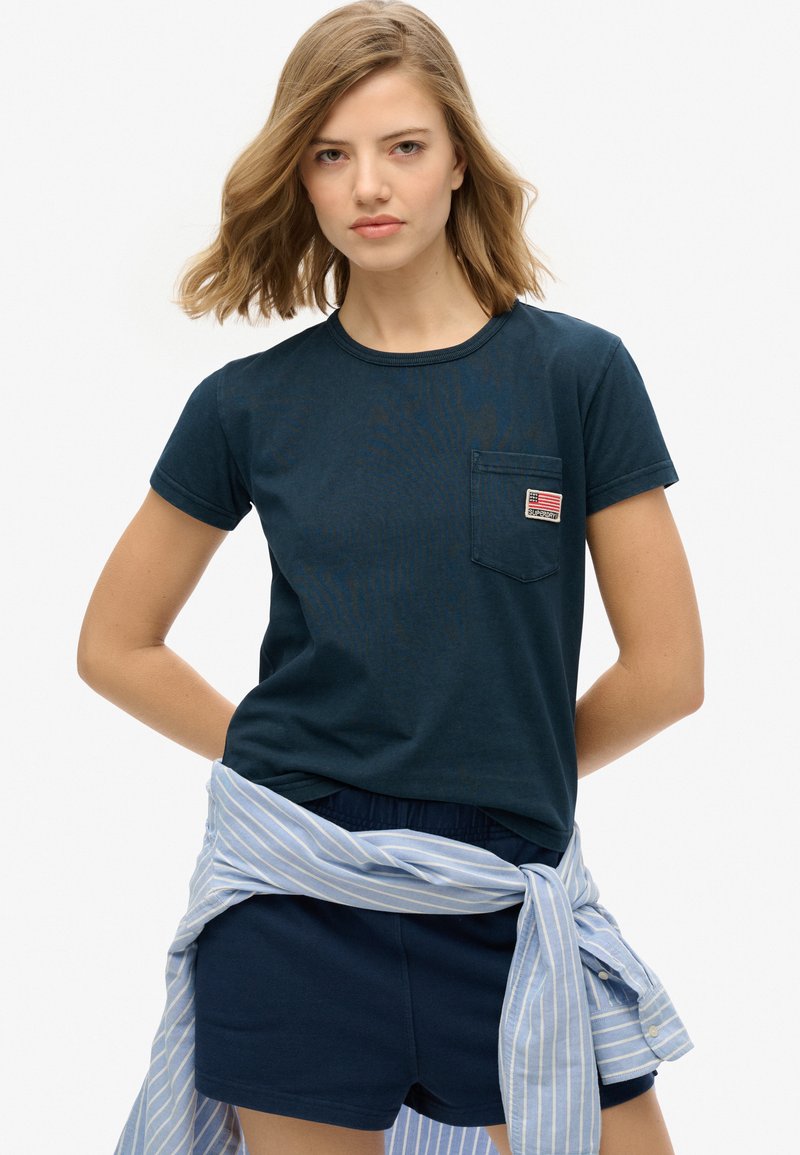 Navyblauwe katoenen t-shirt met korte mouwen, ronde hals en een voorzak met een klein logo-opdruk. Gestyled met navy korte broek en een lichtgestreept shirt om de taille gebonden.