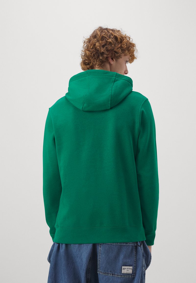 Sudadera verde con capucha y textura suave, con un bolsillo tipo canguro y puños acanalados, combinada con unos pantalones cortos de mezclilla holgados.