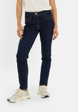 Personne portant un jean slim bleu foncé, des baskets blanches et beiges, et une chemise blanche rentrée, debout devant un fond blanc uni.