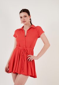 Short-sleeve, button-up dress in coral textured fabric with a fitted waist and flared skirt, featuring a tied belt and a collar.