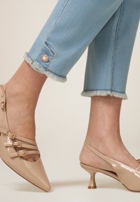 Tacones de charol beige con tira al tobillo, punta afilada y tres correas ajustables. Pantalones de mezclilla cortos en azul claro con bordes desflecados y botón decorativo.