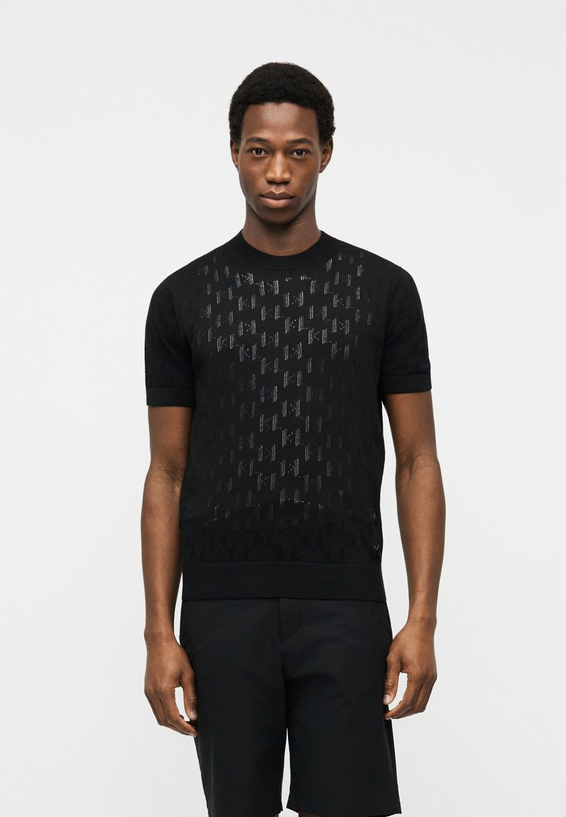 Άνδρας που στέκεται προς τα εμπρός, φορώντας μαύρο πλεκτό πουκάμισο με κοντά μανίκια και διακριτικό μοτίβο, καθώς και μαύρο σορτς, μπροστά σε απλό φωτεινό φόντο.