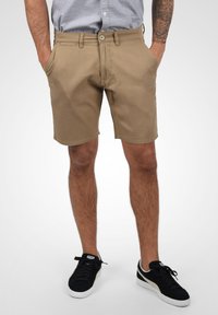 Pantalones cortos de algodón color beige con bolsillos frontales, trabillas y cierre de botón. Se llevan con zapatillas negras, de pie contra un fondo claro.
