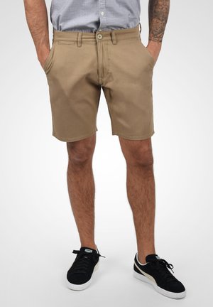 Shorts en coton beige avec poches avant, passants de ceinture et fermeture à bouton. Portés avec des baskets noires, debout contre un fond clair.