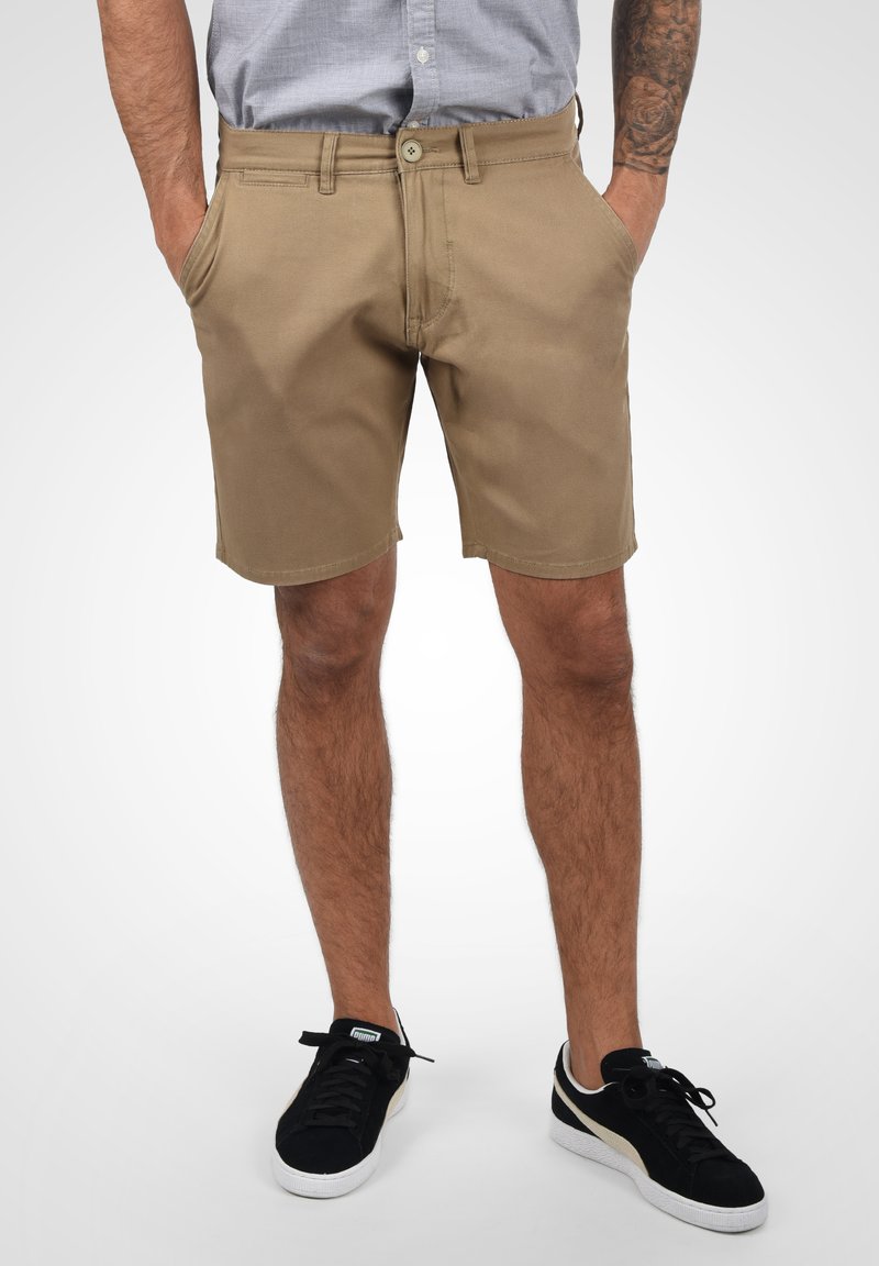 Pantalones cortos de algodón color beige con bolsillos frontales, trabillas y cierre de botón. Se llevan con zapatillas negras, de pie contra un fondo claro.