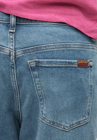 Τζιν παντελόνια σε ανοιχτό μπλε χρώμα, με εμφανή πίσω τσέπη και αντίθετη ραφή. Μια μικρή ετικέτα είναι προσαρτημένη κοντά στην τσέπη.