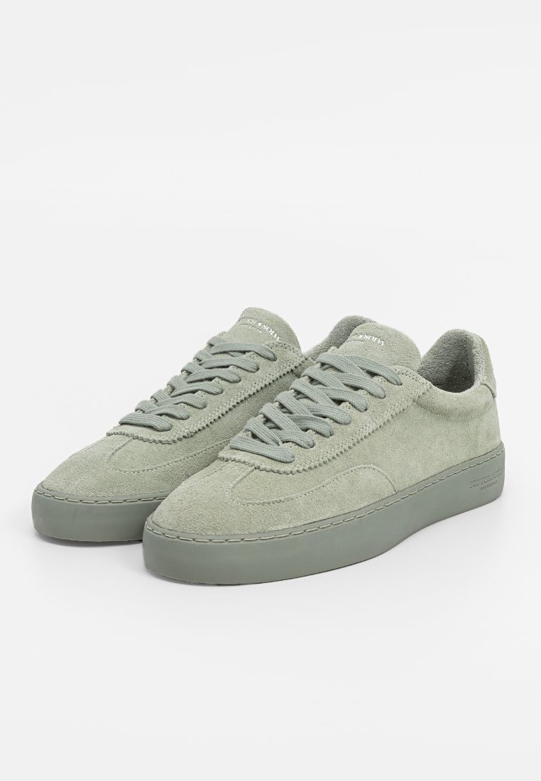 Une paire de baskets basses en daim gris clair avec des lacets assortis et des semelles en caoutchouc épaisses, présentée sur un fond blanc.