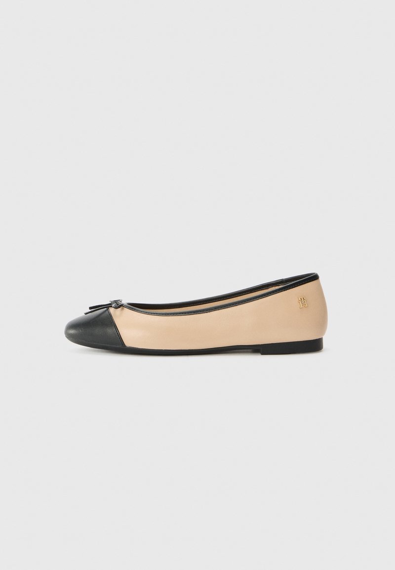 Chaussure de ballet plate beige et noire avec un petit nœud sur le bout et un logo doré près du talon, vue de côté sur un fond uni.