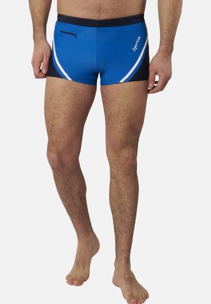 Mann steht barfuß und trägt blaue Badehose mit schwarzem Bund, weißen Streifen und kleinem vorderem Reißverschlusstaschen auf der linken Seite.