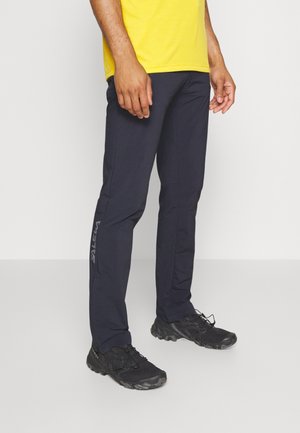 Man wearing black athletic pants and black sneakers standing against a white background, with a yellow shirt partially visible.