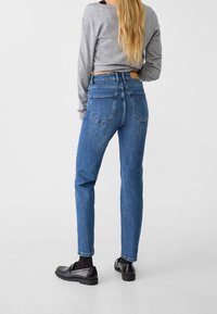 Vatsaa korostavat siniset farkut, valmistettu denimiä, hieman haalistuneet, takataskut ja ruskea nahkainen merkki vyötäröllä. Mustat loafersit jalassa.