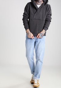 Ciemnoszara bluza z kapturem na zamek, z kieszenią kangurzą, noszona na białej koszulce, w zestawie z jasnoniebieskimi dżinsami i beżowo-białymi trampkami.