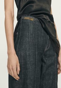 Ciemnoniebieskie dżinsy w szerszym kroju, z ozdobnym złotym guzikiem przy pasie i kontrastowym przeszyciem wzdłuż szwów.