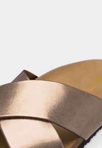 Κοντινή λήψη μπεζ παντόφλας με φαρδιές, υφασμάτινες μεταλλικές λωρίδες σε σχήμα σταυρού και ταμπά μαξιλαρωμένο πέλμα.