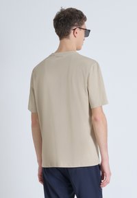 T-shirt beige a maniche corte con scollatura rotonda, texture liscia e un delicato dettaglio del logo vicino al colletto posteriore. Abbinato a pantaloni scuri.