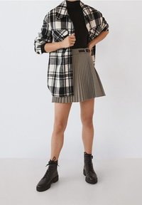 Camisa chaqueta de cuadros en blanco y negro de gran tamaño, combinada con una falda corta de kaki con pliegues y botas de tobillo negras con cordones. Materiales suaves y texturizados.