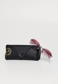 Étui des lunettes de soleil Ray-Ban noir avec un autocollant logo doré et des lunettes de soleil ovales sans monture teintées de violet reposant à côtés sur un fond blanc.
