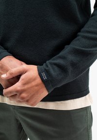 Schwarzer, strukturierter Pullover mit langen Ärmeln, der ein Etikett am Bündchen trägt, über einem hellen Hemd getragen, kombiniert mit olivgrünen Hosen.