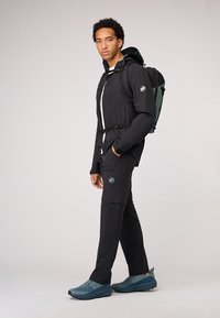 Chaqueta impermeable negra con cierre delantero, acentos grises, pantalones negros y zapatillas deportivas azules. Incluye una mochila verde con correas ajustables.