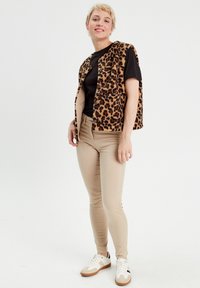 Veste sans manches à motif léopard portée sur un haut noir, associée à un pantalon ajusté beige et des baskets blanches avec des accents noirs.