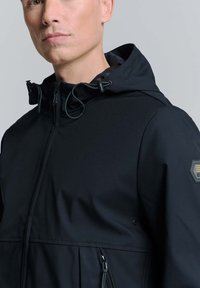 Zwarte hoodie gemaakt van gladde stof, met een ritssluiting, aanpasbare koordjes en een logo patch op de linkerschouder.
