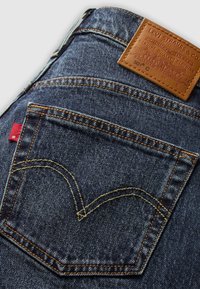Mørkeblå denimbukser med et lærredspatch som har et preget logo. Distinkt baklomme-søm i chevron-mønster, rød merking.