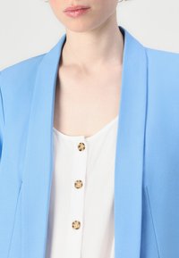 Svetlo modra blazer iz gladkega blaga, z ogrinjalnim ovratnikom. Obučen preko bele srajce z zlatimi gumbi. Čiste linije in krojen fit.