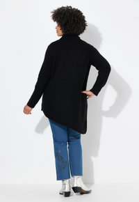 Maglione nero a maglia con colletto alto e orlo inclinato, abbinato a jeans blu con dettagli ricamati e stivali alla caviglia argentati lucidi.
