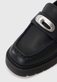 Mocassino in pelle nera con punta arrotondata, dettaglio cucito lungo la parte anteriore e un accento in metallo argentato sulla fascia. Suola testurizzata.