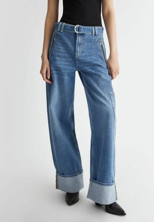 Blauwe jeans met wijde pijpen en omgeslagen zoom, voorzakken met rits, lussen voor een riem en een zilveren D-ring riem, gedragen met zwarte puntige schoenen.