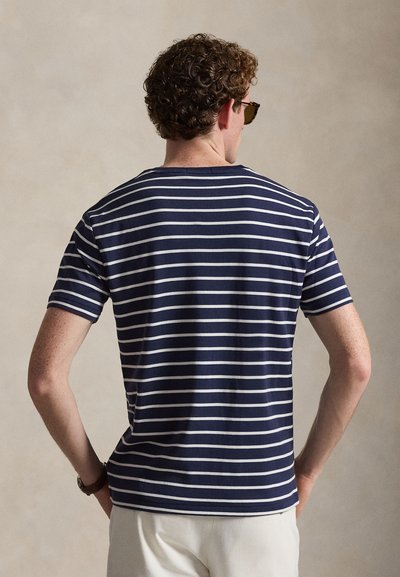 Άνδρας με σγουρά μαλλιά και γυαλιά ηλίου φοράει κοντομάνικο πουκάμισο με ναυτικό μπλε και λευκές ρίγες, στέκεται με τα χέρια στη μέση, στραμμένος προς τα πίσω.