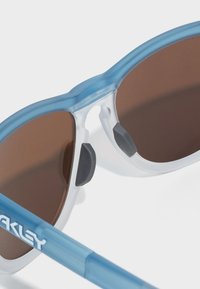 Kék és tiszta napszemüveg barna lencsékkel, textúrázott gumiból készült orrbetétekkel és "Oakley" logóval az ágon. Félkeretes kialakítás.