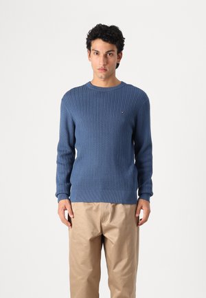 Blauer Strickpullover mit strukturiertem Muster, Rundhalsausschnitt und gerippten Bündchen. Gesticktes Logo auf der Brust; kombiniert mit beigen Hosen.