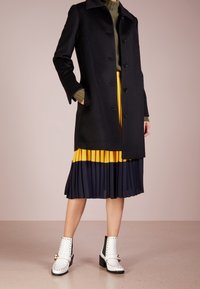 Cappotto di lana nero con colletto e bottoni, abbinato a una gonna plissettata gialla e blu navy e stivaletti bianchi alla caviglia con dettagli neri e borchie.