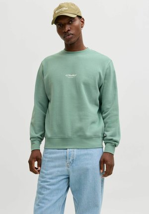 Uomo con felpa verde chiaro JJ Studios, cappellino beige, jeans azzurri, in piedi davanti a uno sfondo chiaro e semplice.