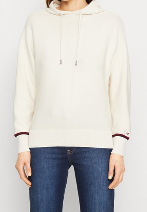 Personne portant un sweat à capuche en maille crème avec cordons et un jean bleu foncé, visible du cou aux hanches, mains détendues le long du corps.