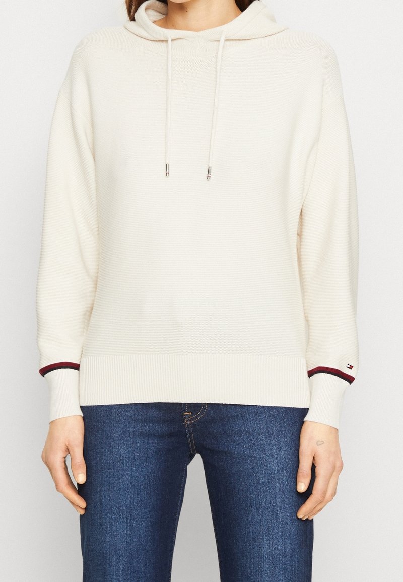 Personne portant un sweat à capuche en maille crème avec cordons et un jean bleu foncé, visible du cou aux hanches, mains détendues le long du corps.