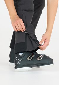 Schwarze Skihosen mit seitlichen Reißverschlüssen, getragen über schwarzen Skischuhen. Die Hosen haben eine leicht strukturierte Oberfläche und einen verstärkten Saum.