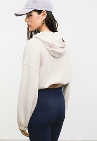 Licht beige cropped hoodie met oversized mouwen en een uitsnijding aan de achterkant; gecombineerd met donkerblauwe hoog-waisted leggings. Grijze pet en hangers.