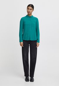 Camicia a maniche lunghe color teal con colletto a camicia e tasca sul petto, abbinata a pantaloni neri. Tessuto morbido, fit rilassato.