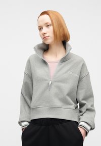 Νεαρό άτομο με ίσια κόκκινα μαλλιά φοράει γκρι φούτερ με φερμουάρ στη μέση και μαύρο παντελόνι, κοιτάζοντας ελαφρώς προς το πλάι μπροστά από λευκό φόντο.