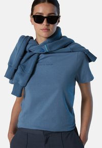 T-shirt di cotone blu con stampa "NORTH SAILS", abbinata a un maglione blu messo sulle spalle, indossato con occhiali da sole neri. Stile casual.