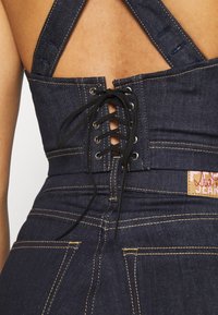 Mörkblå denim jumpsuit med snörning bak, breda axelremmar och kontrasterande gul sömnad. Har en märkesetikett vid midjan.