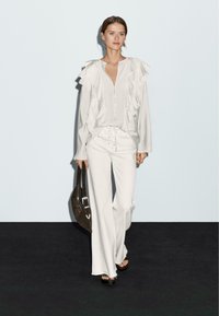 Vrouw in een witte, geplooide blouse en veterbroek met brede pijpen, met een donkeren bruine tas, staande op een zwarte vloer tegen een witte muur.