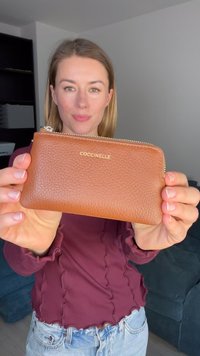 Portefeuille en cuir marron avec une texture en relief, logo doré "Coccinelle", fermeture éclair, forme rectangulaire et design compact.