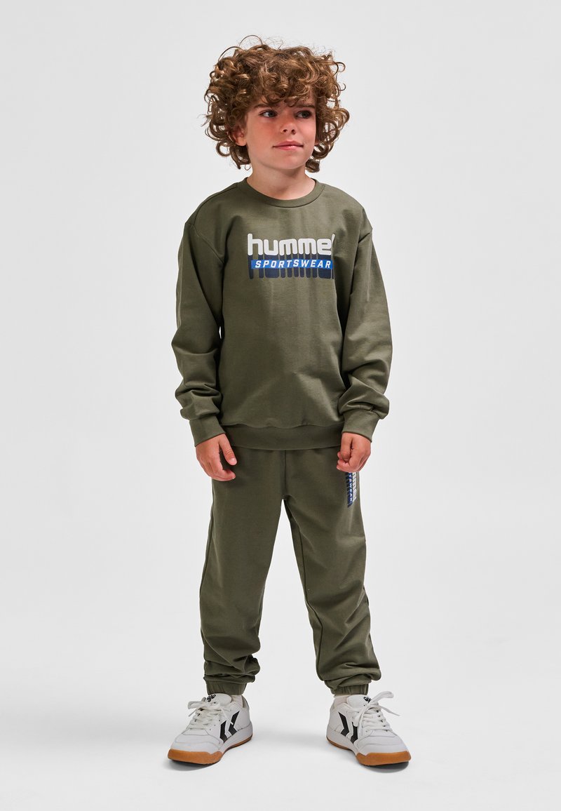Olivengroen sweatshirt met het "hummel SPORTSWEAR" logo, gecombineerd met bijpassende joggingbroek en witte sportschoenen met zwarte accenten.