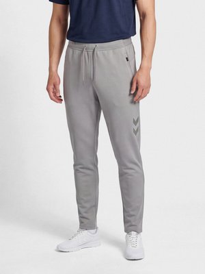 Persona con pantalones deportivos grises ajustados con bolsillos laterales y zapatillas blancas, de pie frente a un fondo claro y sencillo.