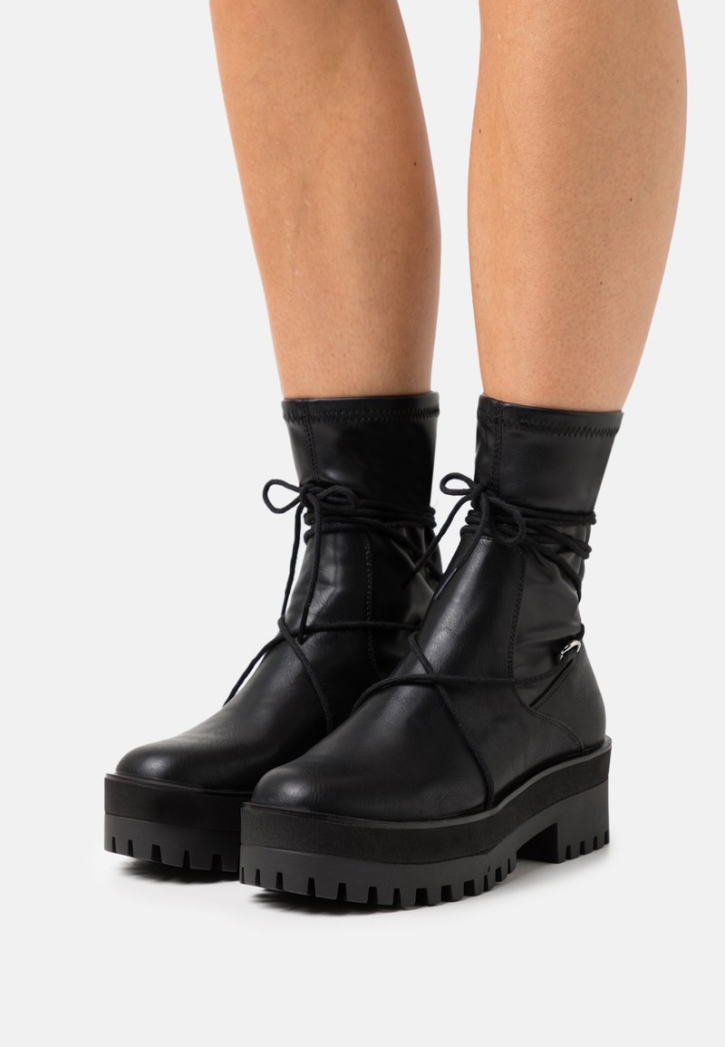Bottines à plateforme en cuir noir avec semelles épaisses à crampons et lacets enroulés portées sur jambes nues devant un fond blanc.